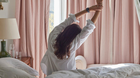 Woman stretching in bed beside a sunlit window, highlighting the importance of rest, recovery and starting the day feeling refreshed.