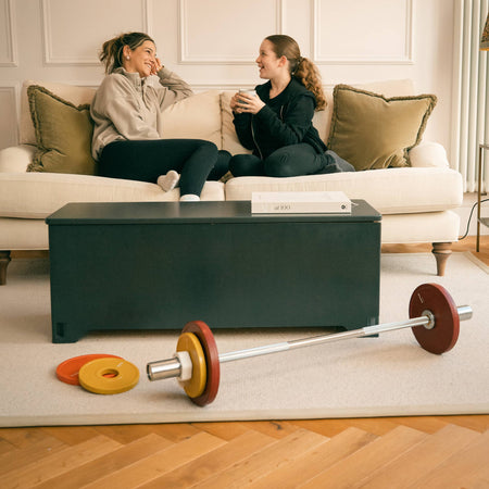 Two women sitting on a couch in a living room with a midnight Fittle box acting as a coffee table and a Fittle barbell and weight plates loaded in the foreground.