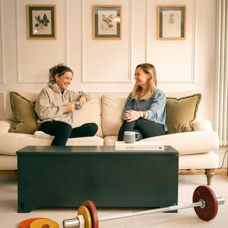 Two women sitting on a sofa in a modern living room with a midnight Fittle Box home gym storage bench used as a coffee table, barbell and weight plates on the floor, showcasing stylish design-led home strength training equipment in a UK home interior.