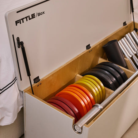 A soft grey Fittle Box with its lid open, showing the max pack, which includes 4 10kg charcoal weight plates. The box is positioned next to a bed with white bedding.