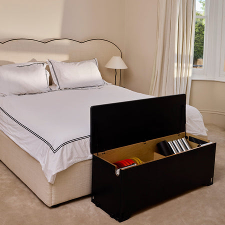 A midnight Fittle box with its lid open, revealing storage compartments with colorful items inside, positioned at the foot of a neatly made bed in a warm beige bedroom.