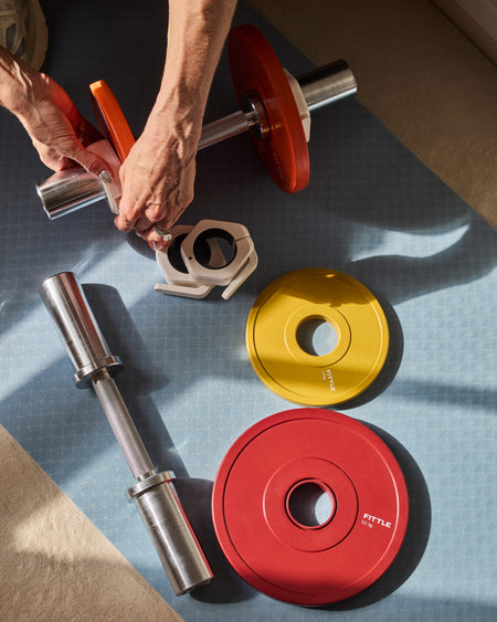 A top-down view of a person's hands assembling a Fittle adjustable dumbbell with red and orange weight plates on a blue textured mat.