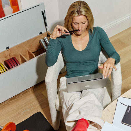 A woman sits in a white armchair in a home office environment, wearing a teal long-sleeve top, cream pants, and red socks while working on a laptop. A Fittle Box is in the background with a Fittle dumbbell resting on a black mat on the wooden floor nearby.