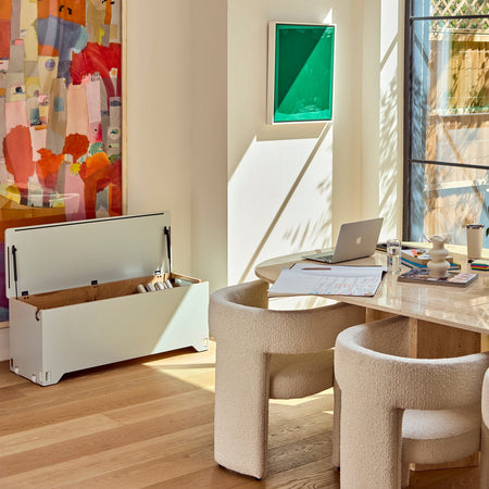 A bright, modern interior corner with warm sunlight streaming in from a large window. The room features a light wood desk, two cream boucle chairs, a colorful abstract painting on the left, a small green framed artwork on the wall, and an open soft grey Fittle Box on the floor.
