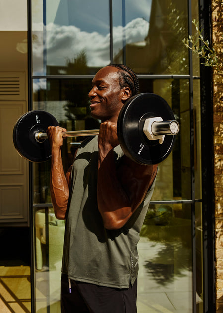 A man lifting a Fittle barbell with black weight plates in front of a glass building wall, wearing a dark olive green sleeveless shirt and black shorts.