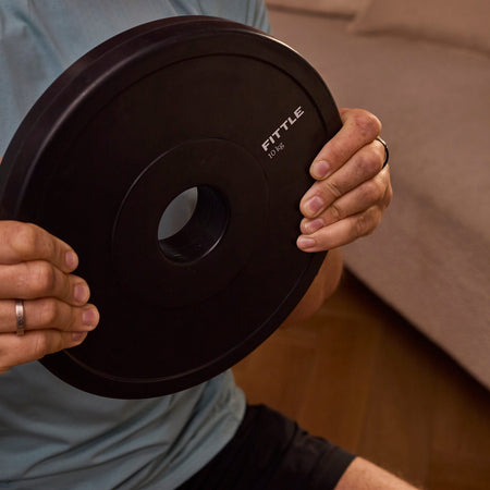 A man holding a black rubber 10kg Fittle weight plate. The man is wearing a light blue-gray t-shirt and black shorts, with a beige couch and wooden floor in the background.