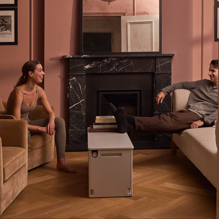 A modern living room scene with a man and a woman seated on opposite sides of a fireplace, with a soft grey Fittle box placed on the floor between them. The room has dusty rose walls, wood flooring, a black marble fireplace, and a large mirror above it.