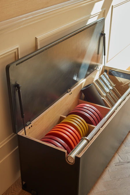 A midnight Fittle Box with its lid open, revealing a wooden interior organized with colorful weight plates in red, orange, and yellow, a steel barbell, dumbbells, and a charcoal mat. 