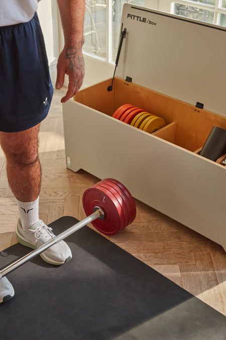 Man in athletic clothing standing next to an open Fittle box with a barbell.