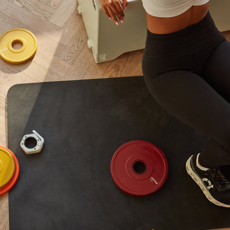 Woman working out surrounded by Fittle Weights on a black mat with wood flooring. 