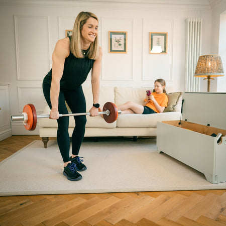 Woman lifting Fittle adjustable barbell for bent over row while child relaxes on sofa in home gym