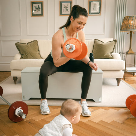Woman performing seated dumbbell curl with Fittle adjustable dumbbells while baby sits on floor nearby