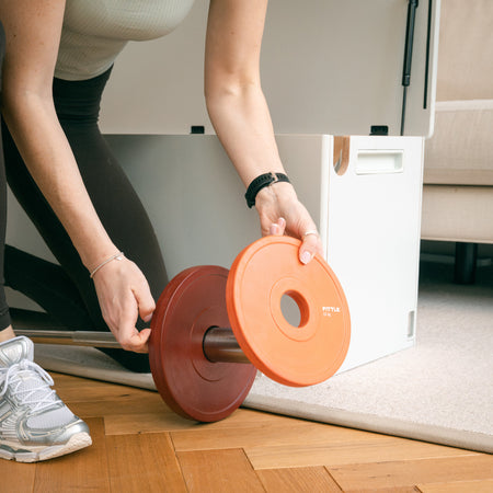 Woman adding Fittle weight plate to adjustable barbell beside the Fittle home gym storage bench
