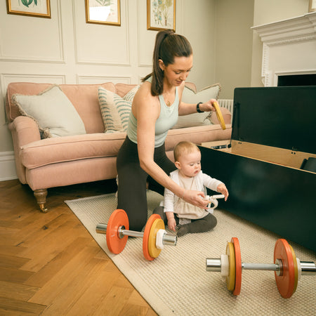 Woman setting up Fittle adjustable dumbbells beside open Fittle Box home gym while child sits nearby in living room