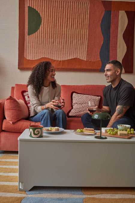 Closed grey Fittle Box used as a coffee table in a modern living room as two people enjoy drinks, showing discreet multifunctional home strength equipment.