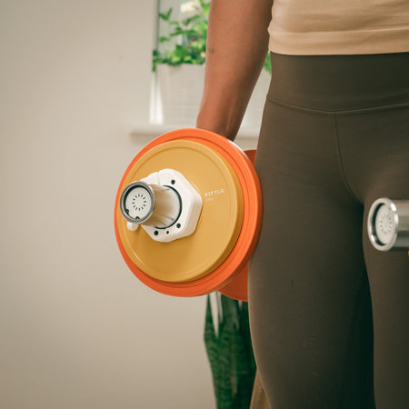 Person holding an adjustable Fittle dumbbell with yellow and orange plates, secured by white clips. 