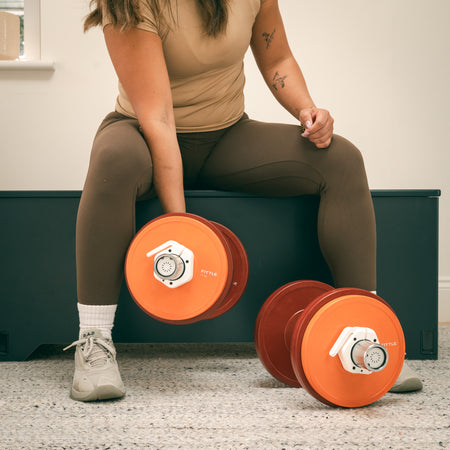 Person sitting on a box performing bicep curls with adjustable Fittle dumbbells in a home setting.