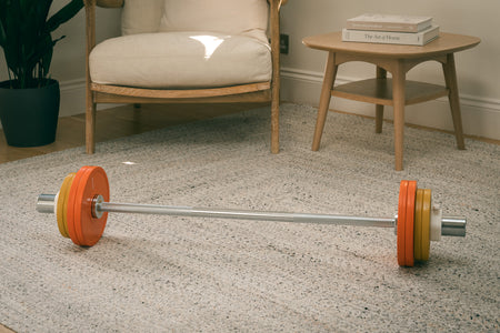 Fittle barbell with orange and yellow weight plates on a carpeted floor in a room with furniture.