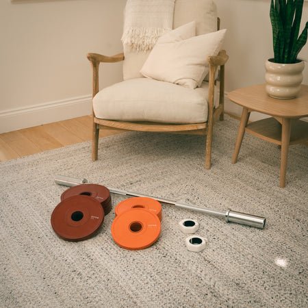 Fittle Barbell with red and orange weights on a carpeted floor in a living room setting.